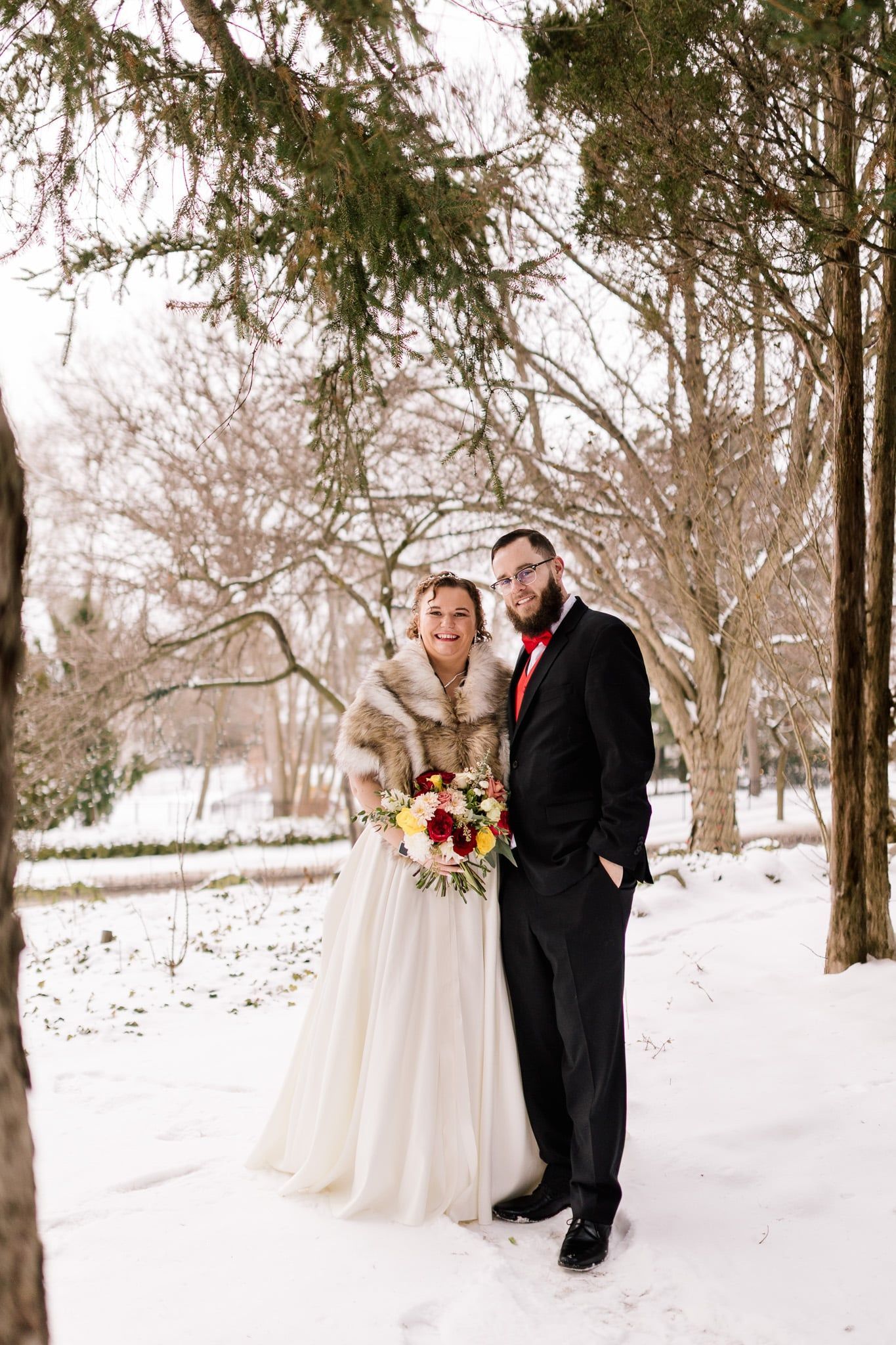 Bride and groom in wedding attire pose in a snowy park setting. The bride wears a fur shawl.