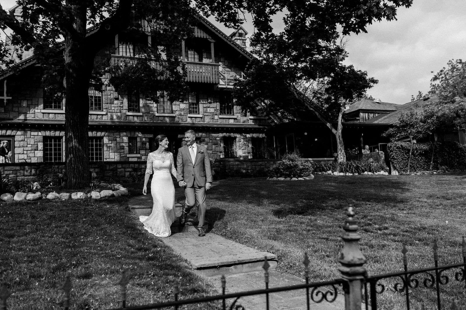 A black and white photo of a bride and groom walking in front of a house.