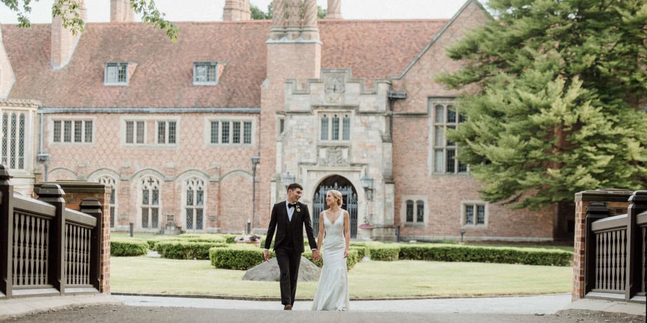 A bride and groom are walking in front of a large brick building holding hands.
