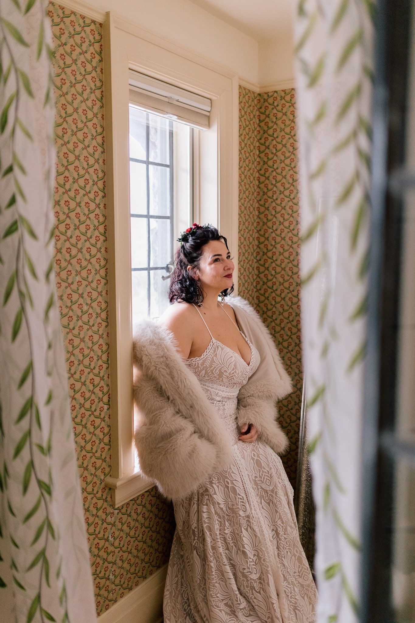 A woman in a wedding dress and fur coat is standing in front of a window.