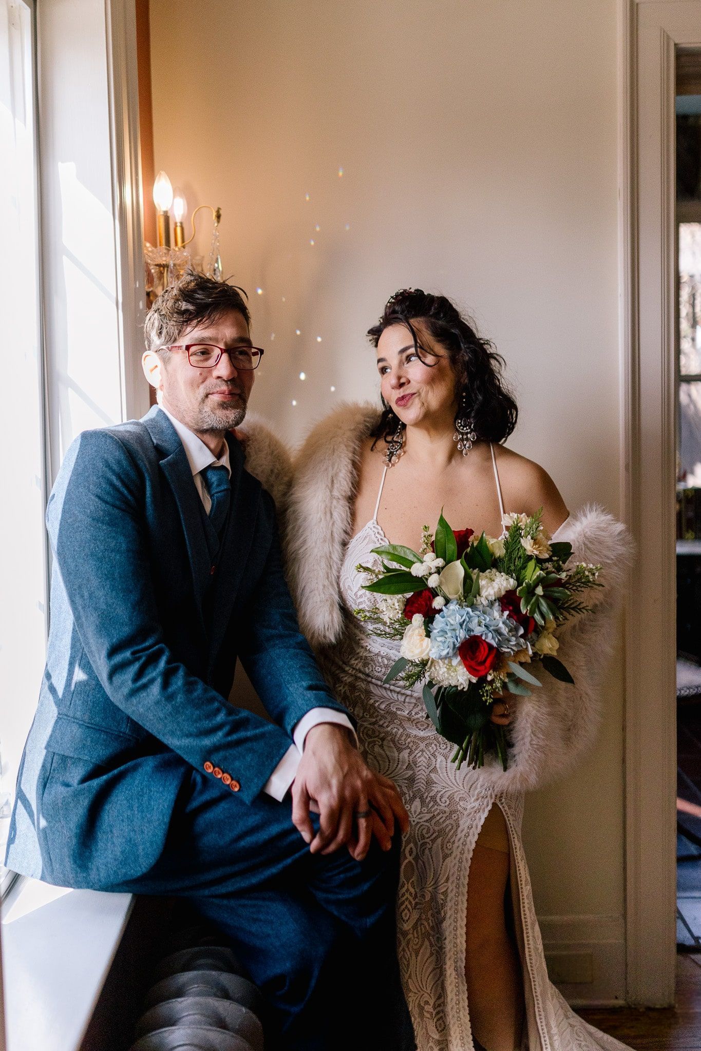 A bride and groom are sitting on a window sill holding hands.