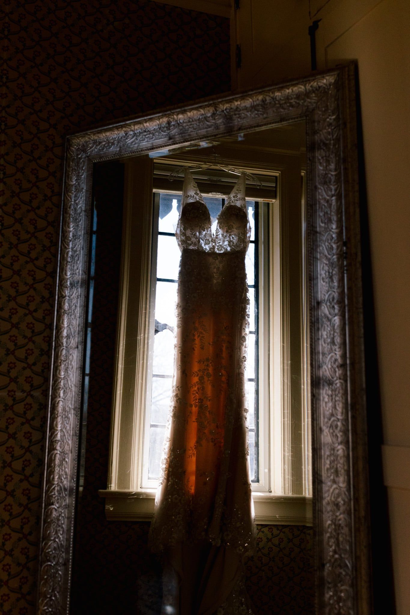 A wedding dress is hanging in front of a window in a mirror.