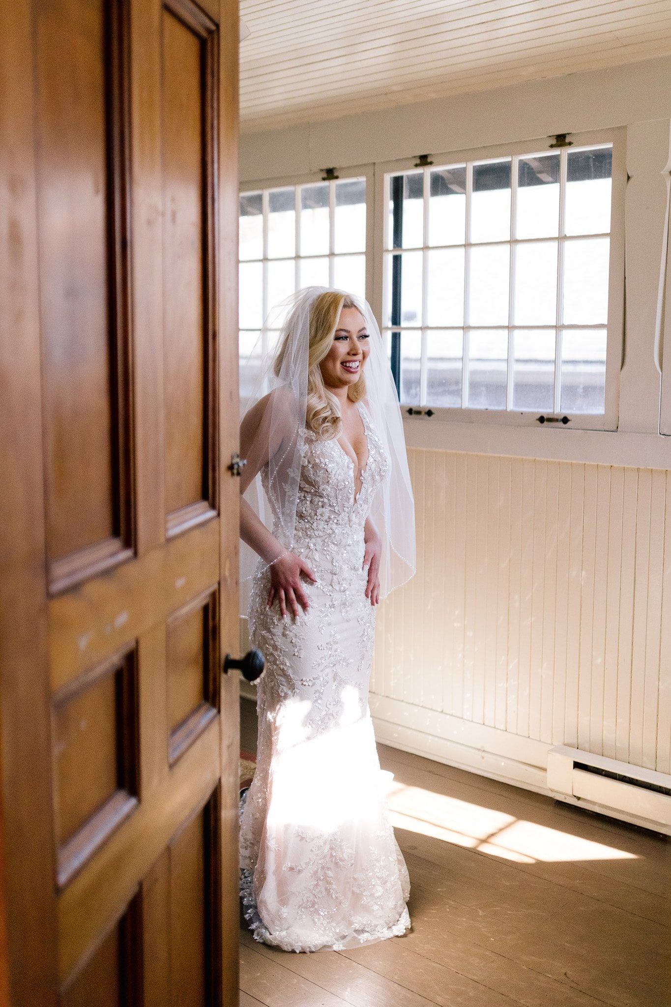 A bride in a wedding dress and veil is standing in a room next to a window.