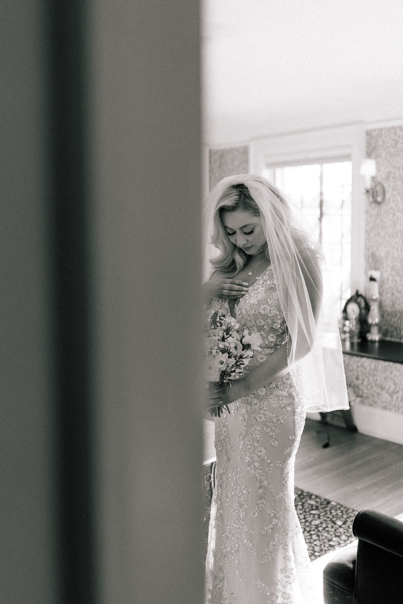 A bride in a wedding dress is standing in a bathroom holding a bouquet of flowers.