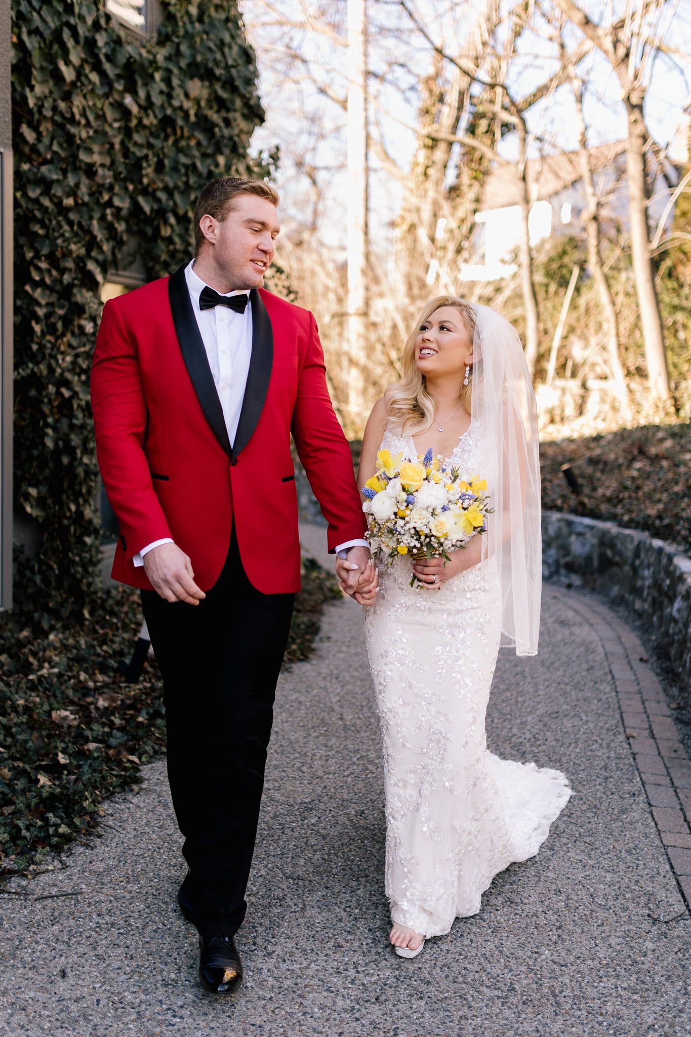 A bride and groom are walking down a sidewalk holding hands . the groom is wearing a red tuxedo.