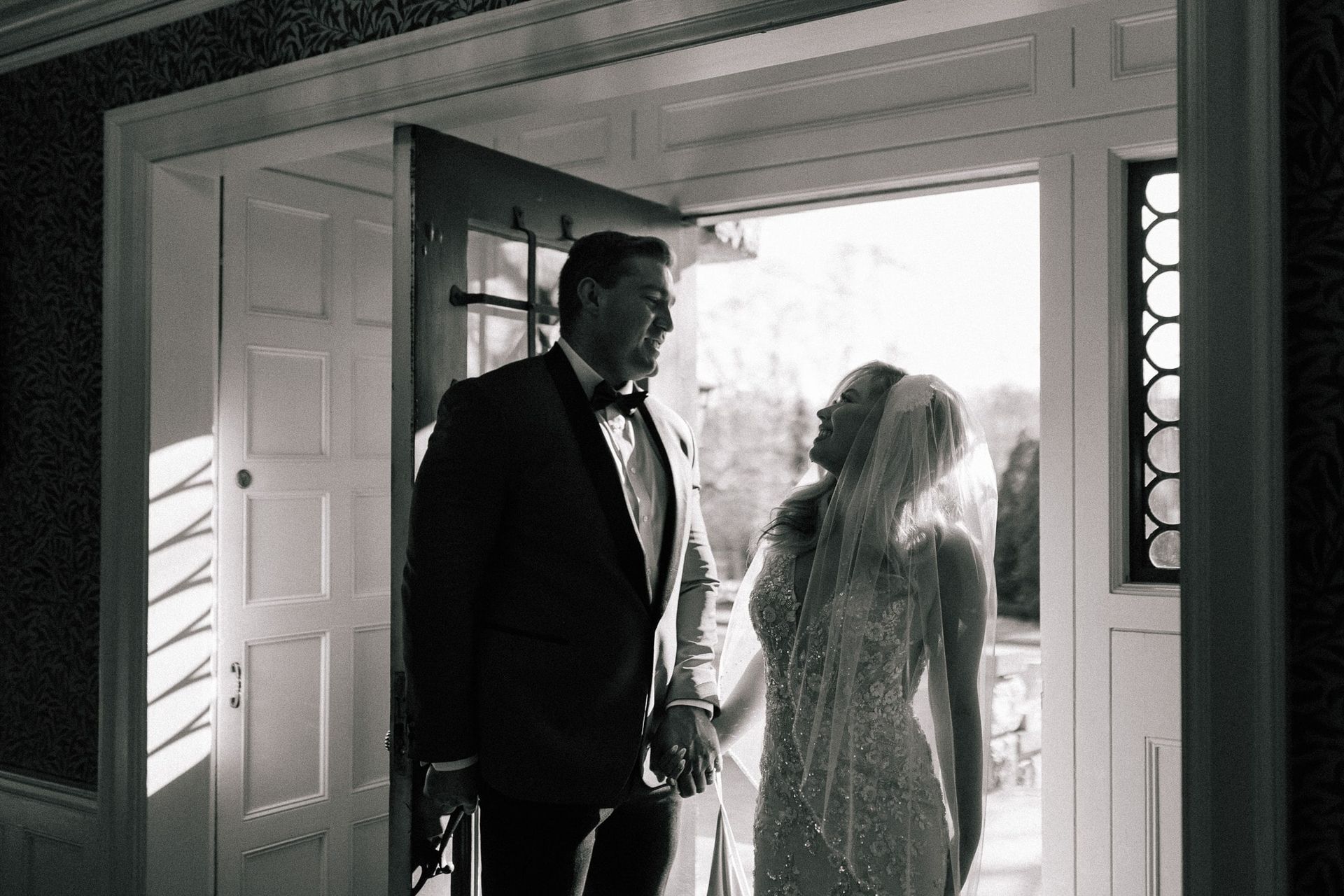 A black and white photo of a bride and groom holding hands in a doorway.