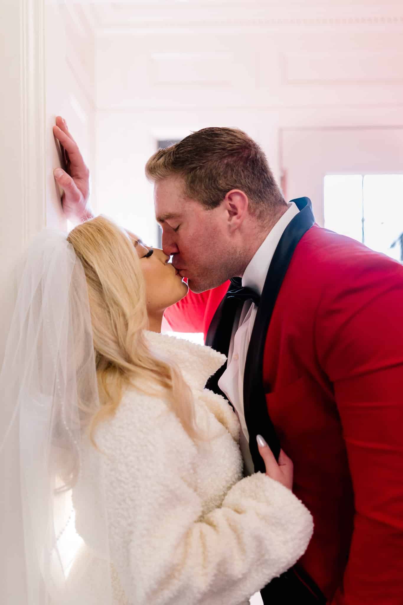 A bride and groom are kissing while leaning against a wall.