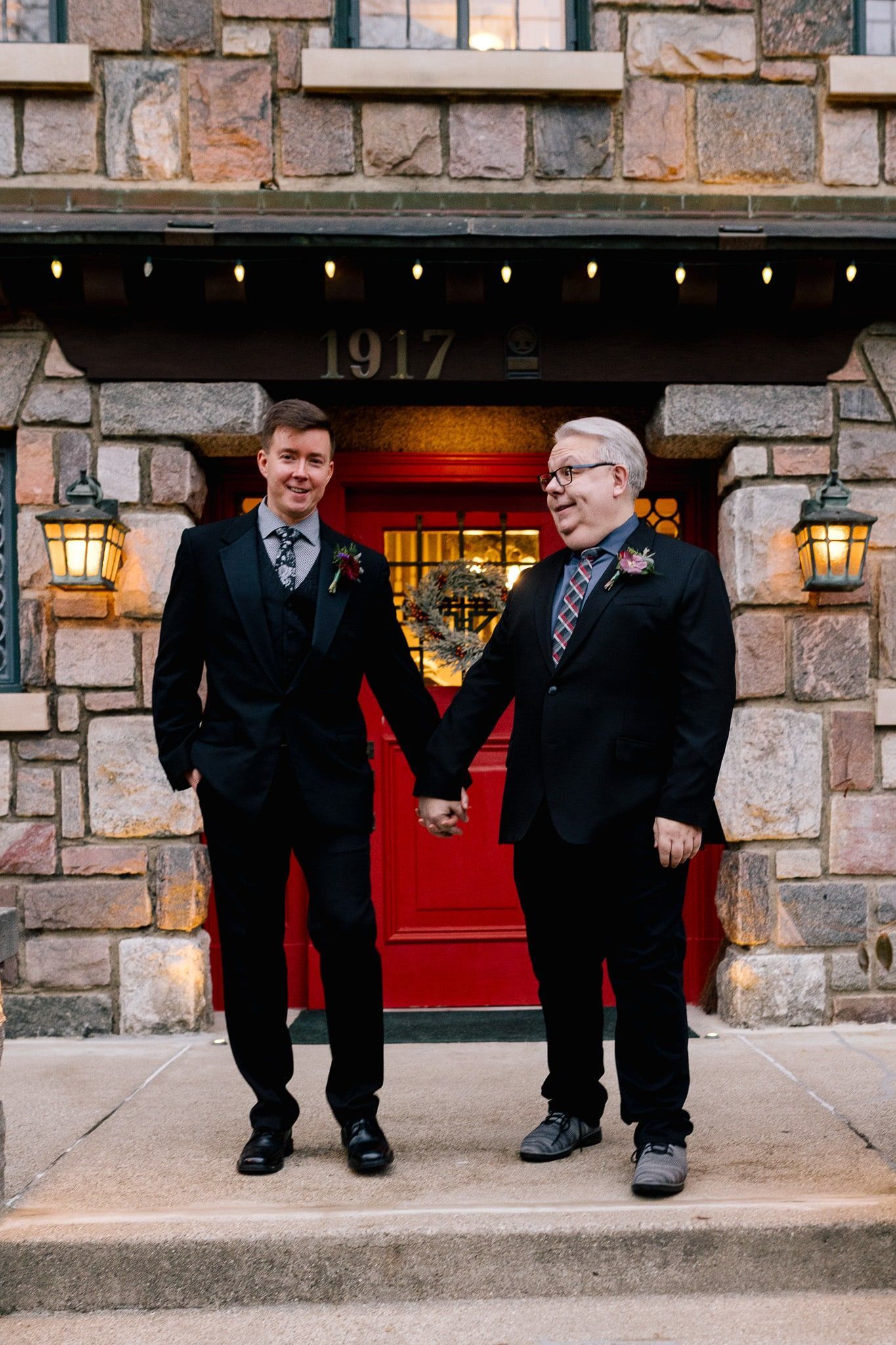 Two men in suits are holding hands in front of a red door.