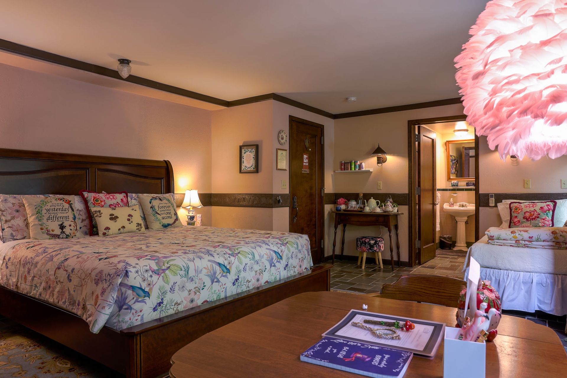A bedroom with a large bed , a table , and a pink feather lamp.