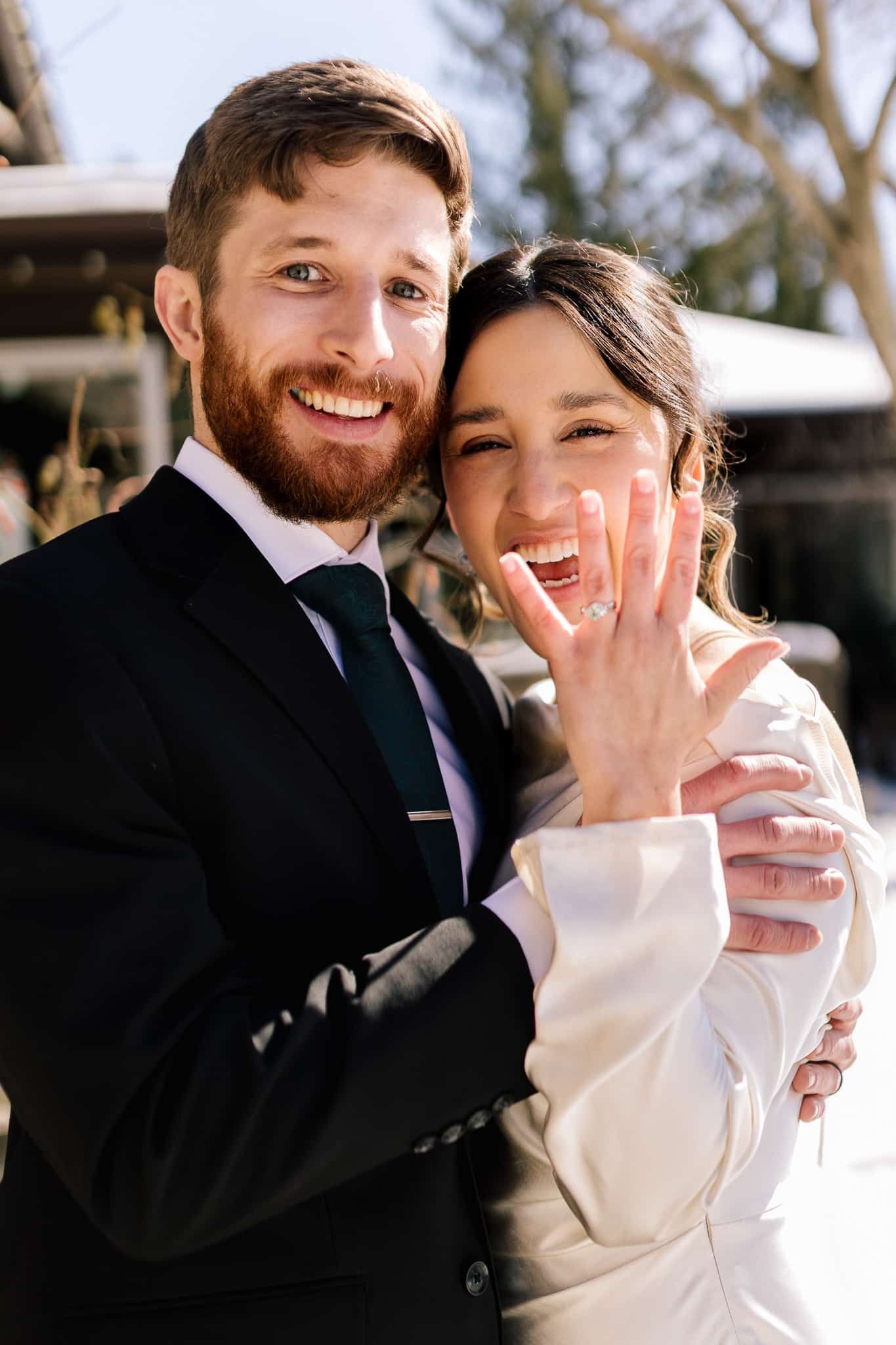 Smiling couple, bride holding up hand showing off ring, groom embracing her, sunny outdoor setting.