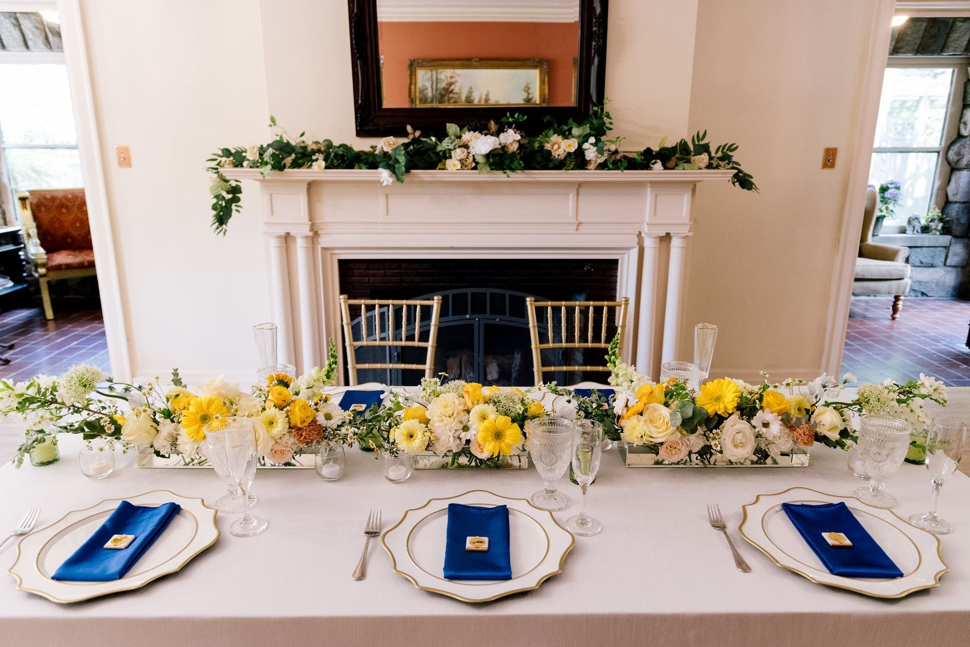A long table with plates , napkins , glasses and flowers in front of a fireplace.