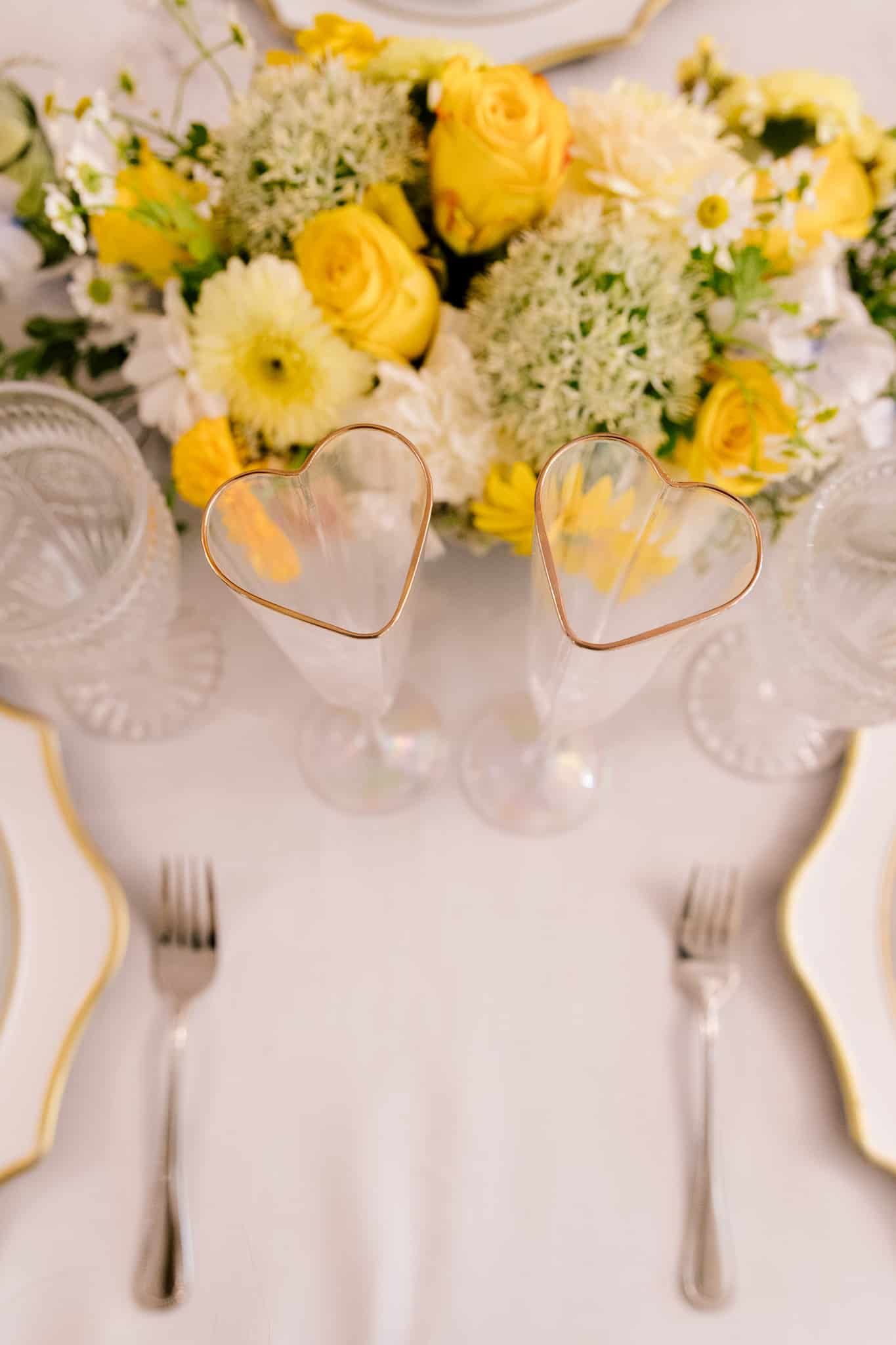 A table with a vase of yellow flowers and two heart shaped wine glasses.