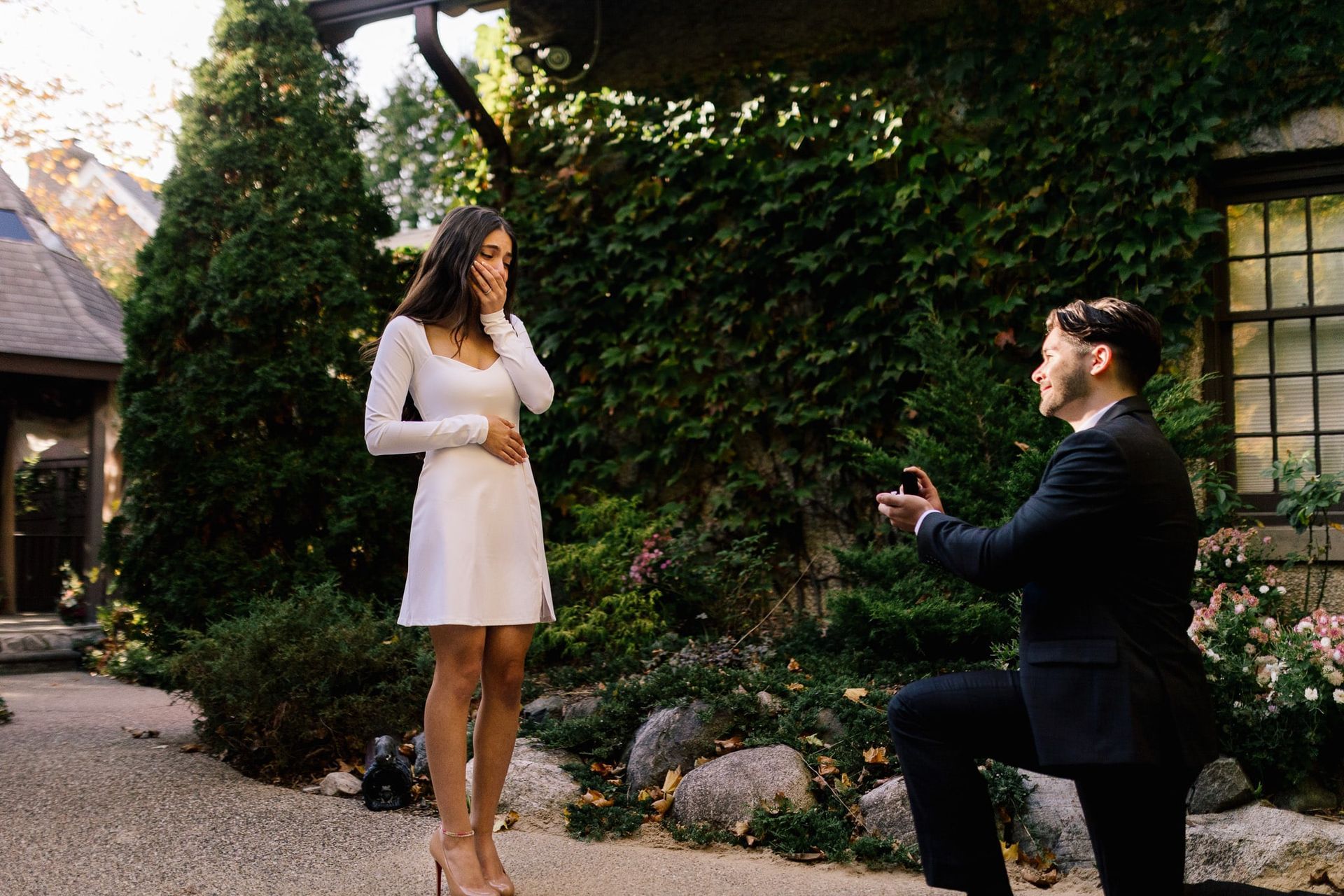 A man is kneeling down to propose to a woman in a white dress.