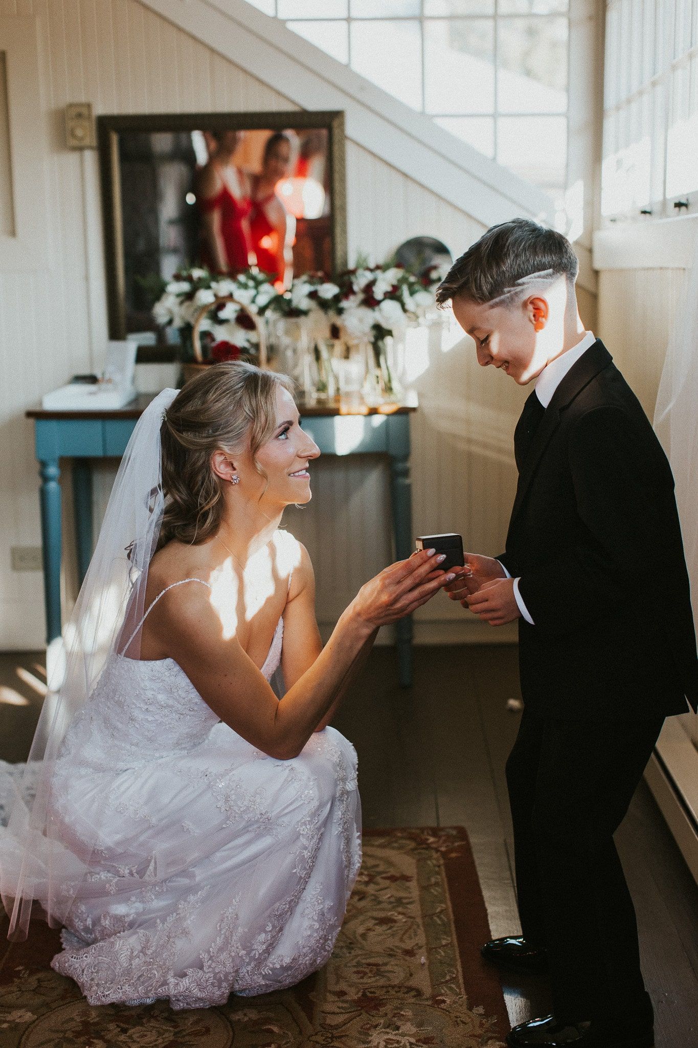 A bride in a wedding dress is kneeling down next to a boy in a tuxedo.