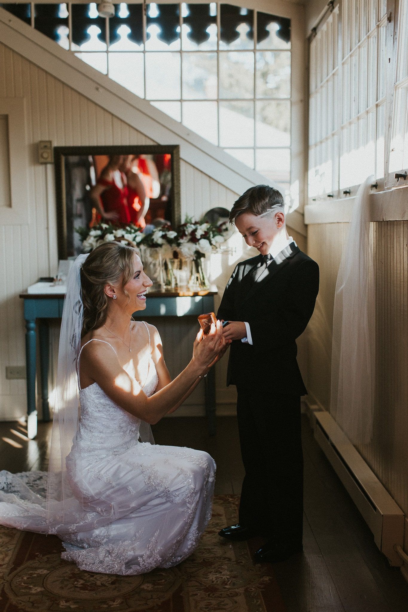 A bride is kneeling down next to a boy in a tuxedo.