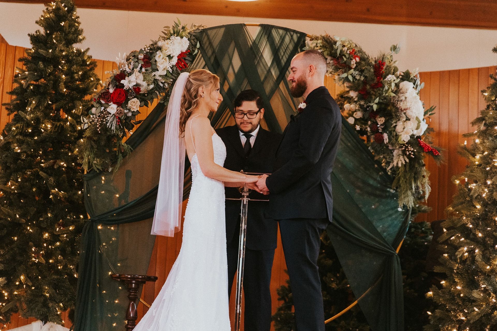 A bride and groom are holding hands during their wedding ceremony in front of christmas trees.