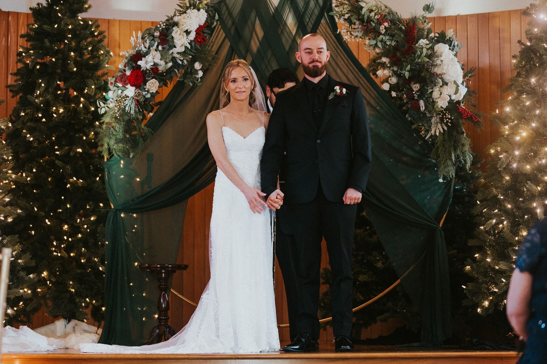 Bride and groom holding hands during a wedding ceremony. They stand before a green draped archway and Christmas trees.
