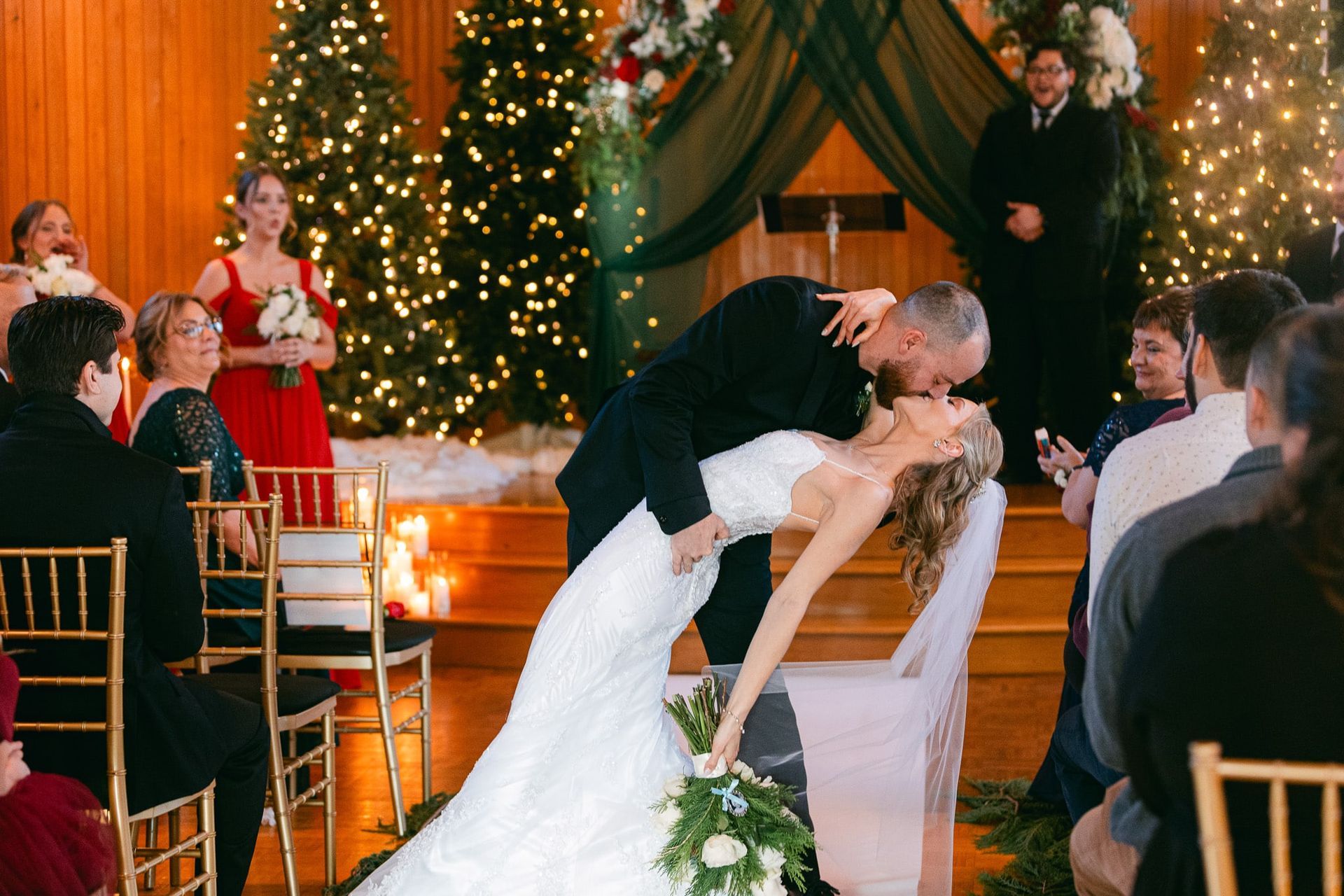 Couple kissing, bride leaning back, at a wedding ceremony decorated with Christmas trees and lights.