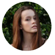 Young woman with long auburn hair looks directly at the viewer, set against a blurred green foliage background.