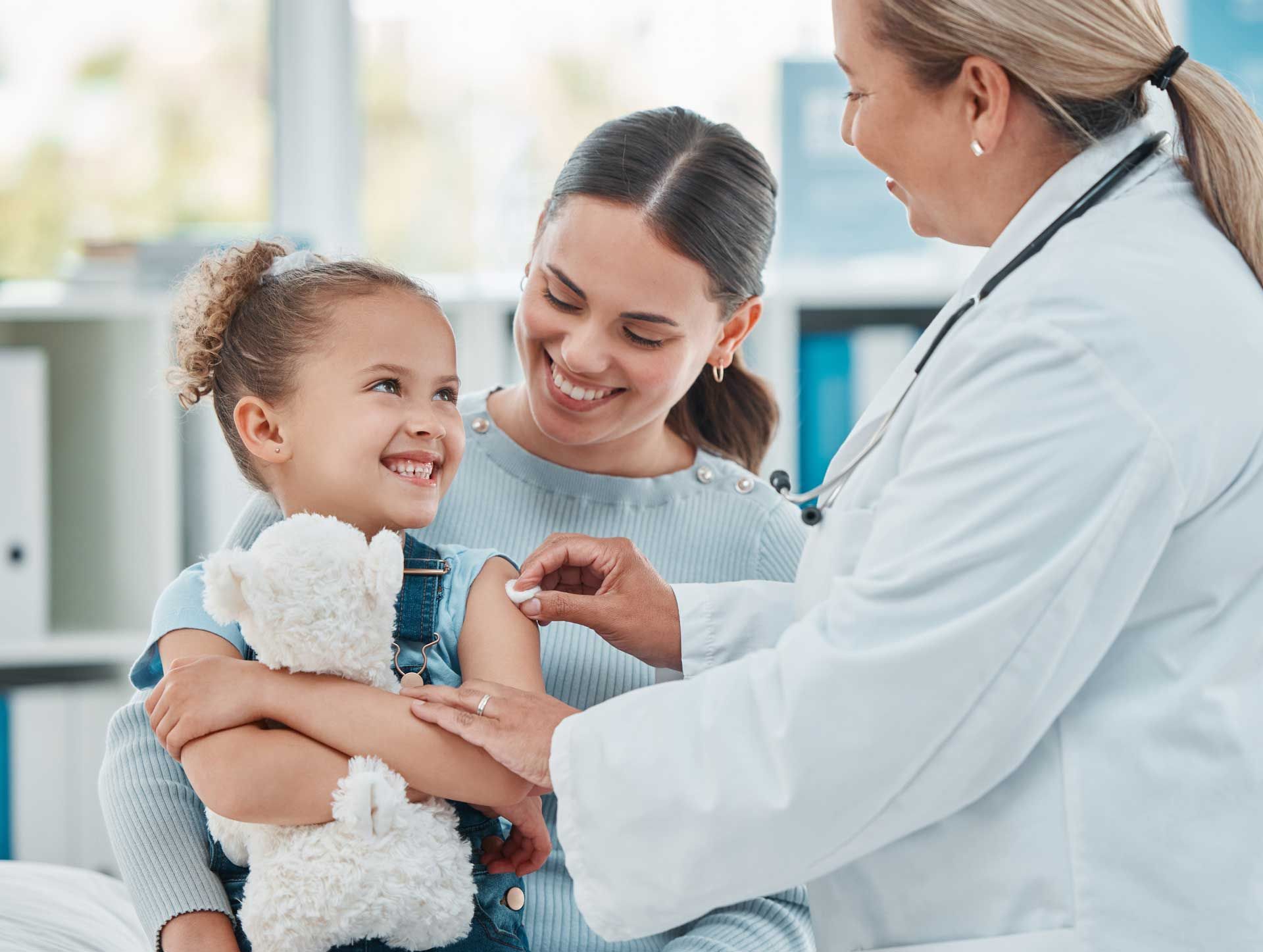 A little girl is getting a vaccine from a doctor while holding a teddy bear.