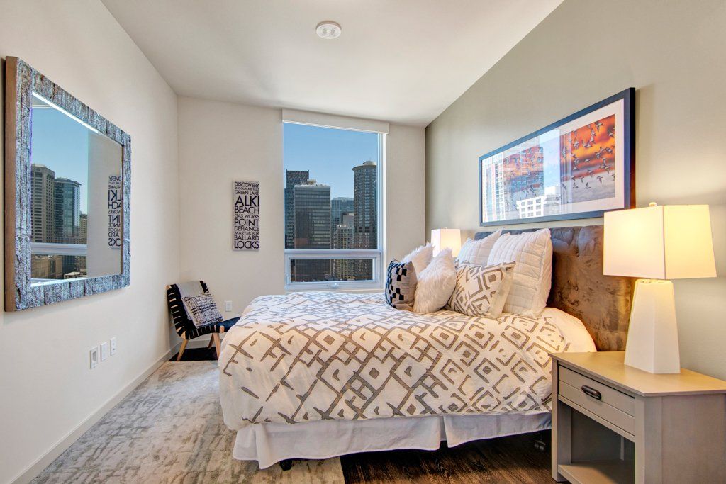 A bedroom with a large bed, nightstand, lamps, and a large mirror at Tower12 Apartments.