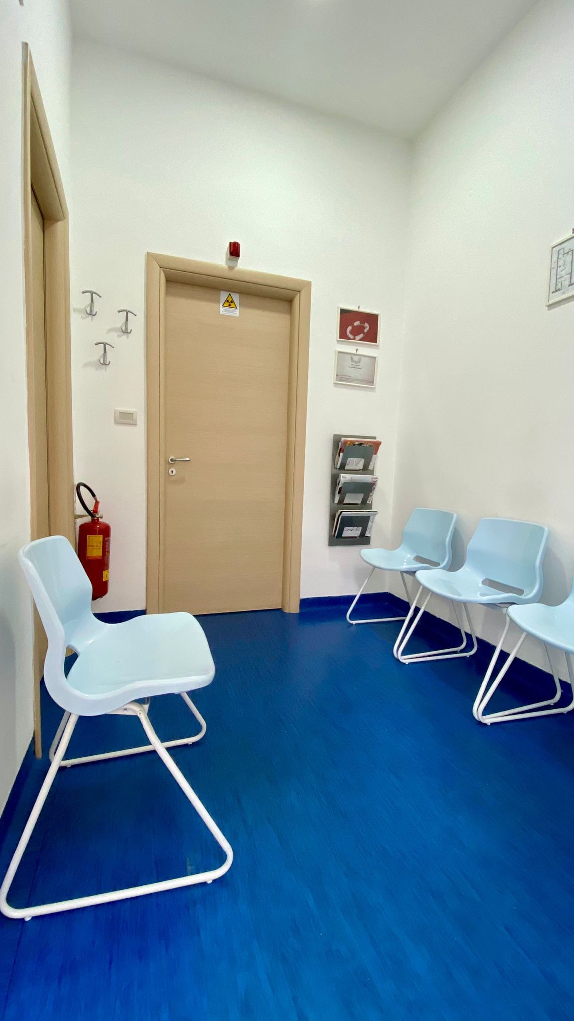 Sala d'attesa con sedie blu, pavimento e porta chiusa. Estintore a sinistra.