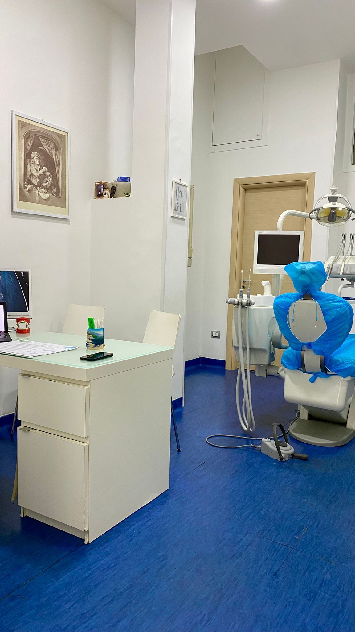 Interno di uno studio dentistico con pavimento blu, scrivania bianca, poltrona odontoiatrica e attrezzatura.