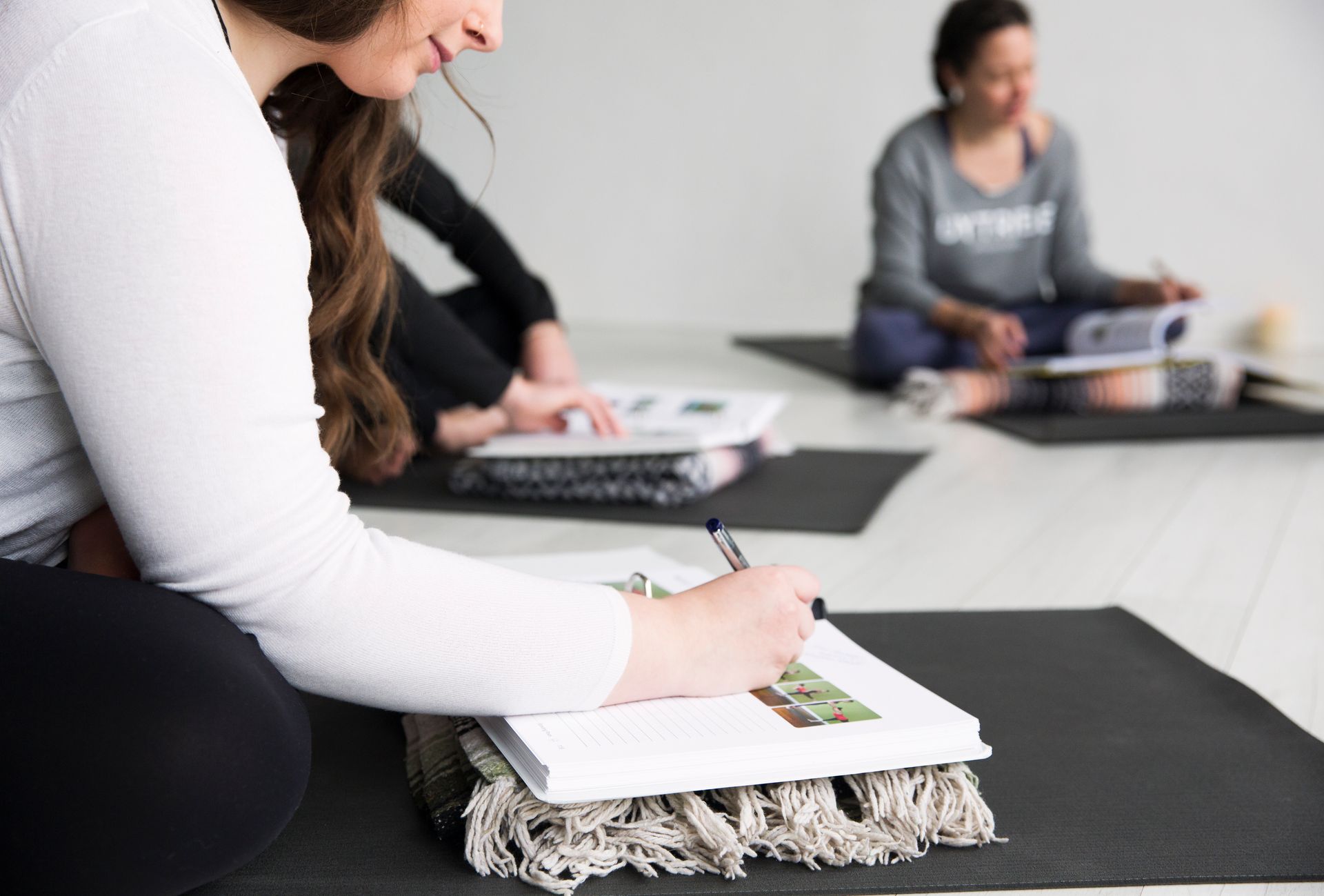 Yoga trainee taking written notes in a 250-page curriculum manual during an in-studio lecture session.