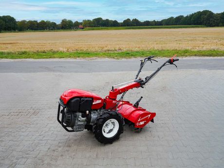 Rote Motorhacke auf grau gepflastertem Untergrund, im Hintergrund ein Feld und Bäume.