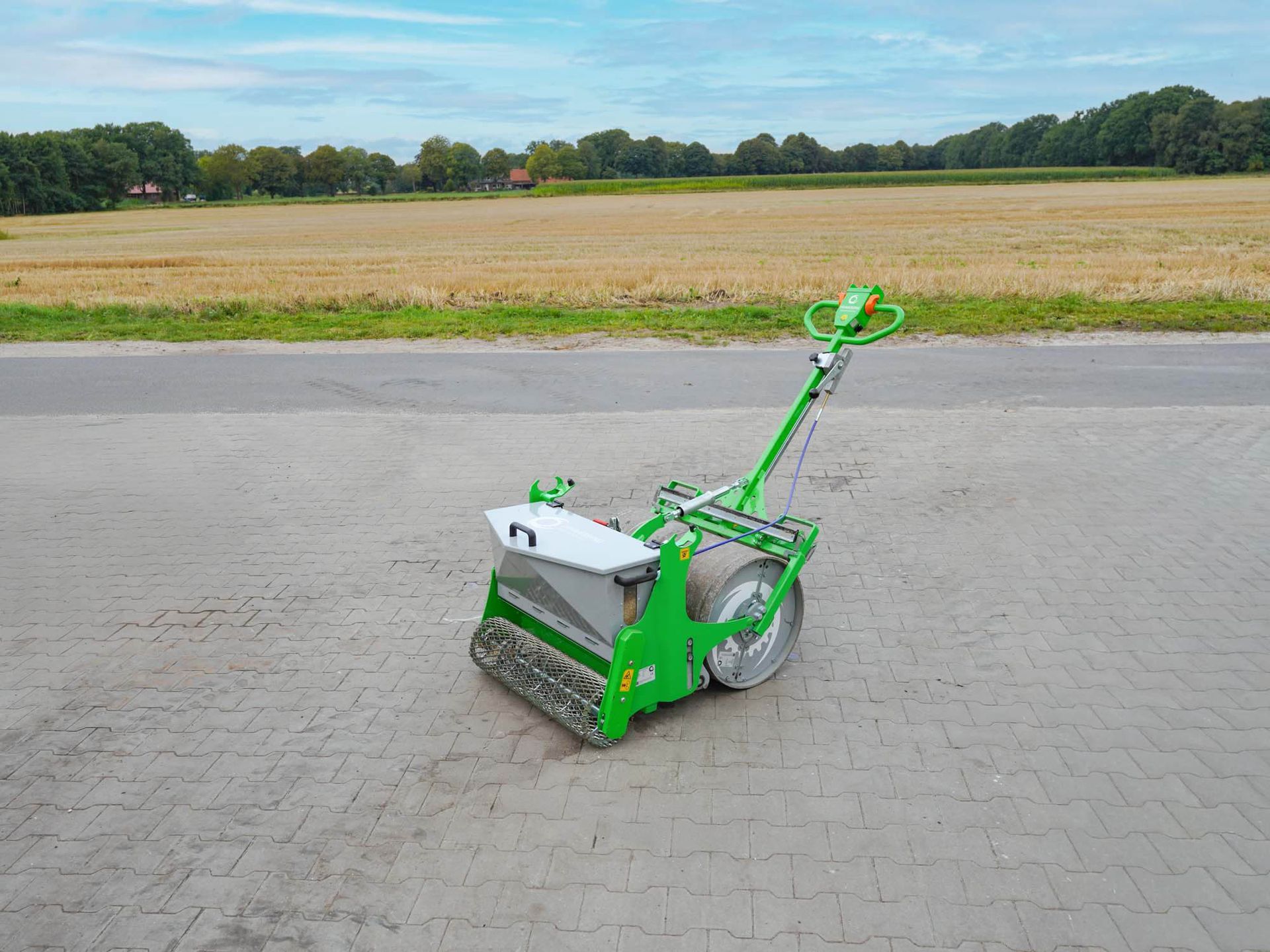 Grün-silberne Feinsaat-Sämaschine auf einer gepflasterten Fläche; Feld und Bäume im Hintergrund.