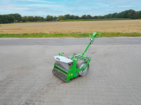 Grün-silberne Feinsaat-Sämaschine auf einer gepflasterten Fläche; Feld und Bäume im Hintergrund.