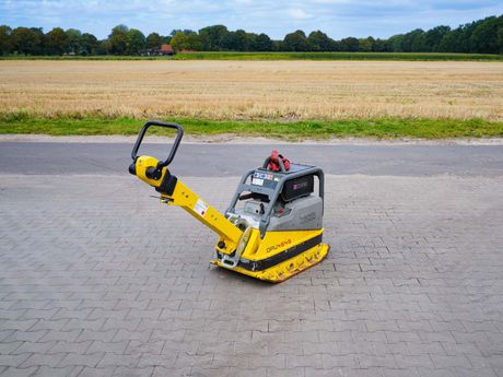 Gelbe und schwarze Rüttelplatte auf einer gepflasterten Fläche, Feld im Hintergrund.