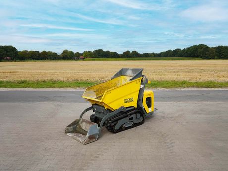 Gelber Raupenkipper mit Hochbett, auf einer asphaltierten Straße, Feld im Hintergrund.