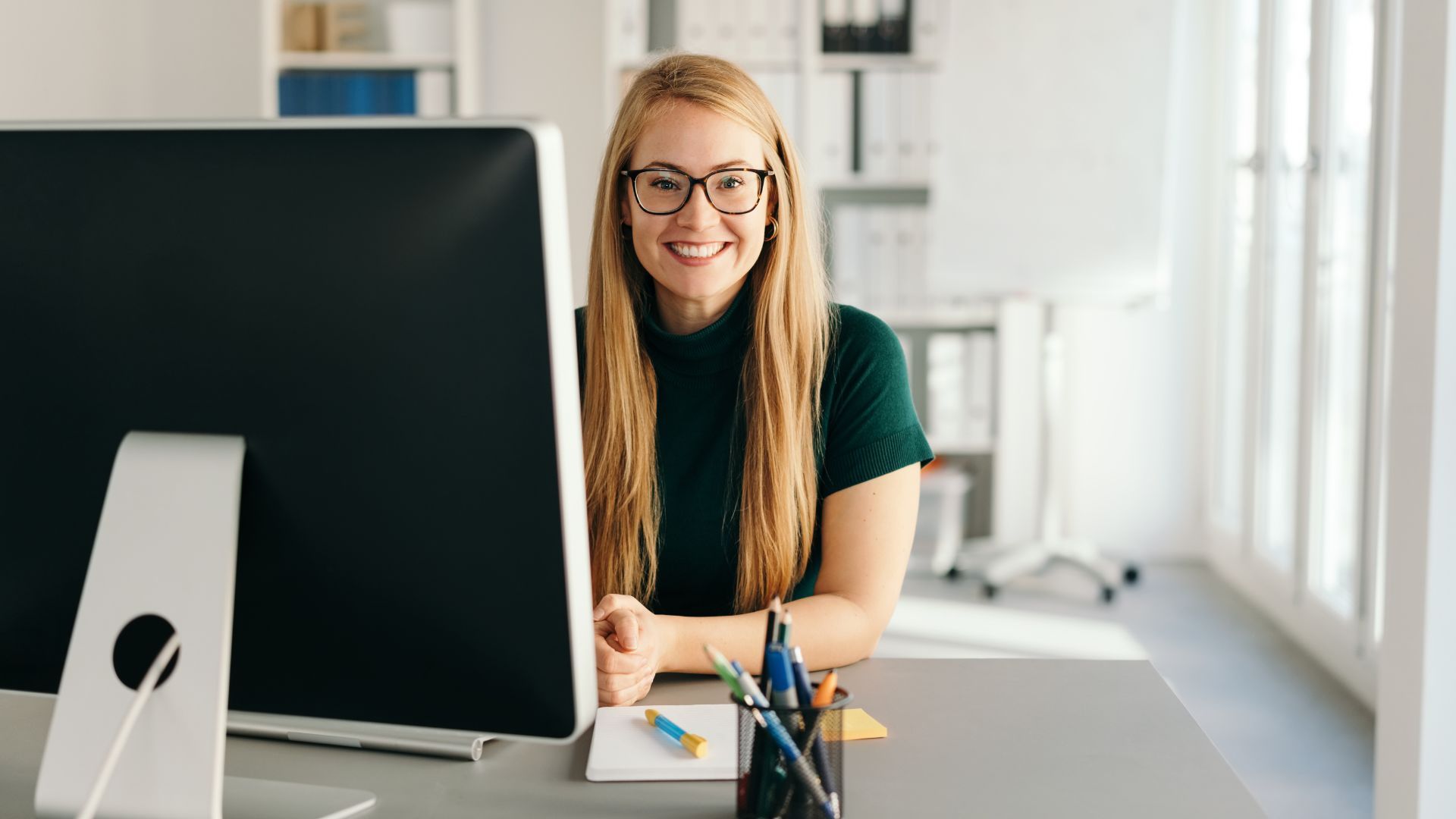 A woman wearing glasses is sitting at a desk in front of a computer.