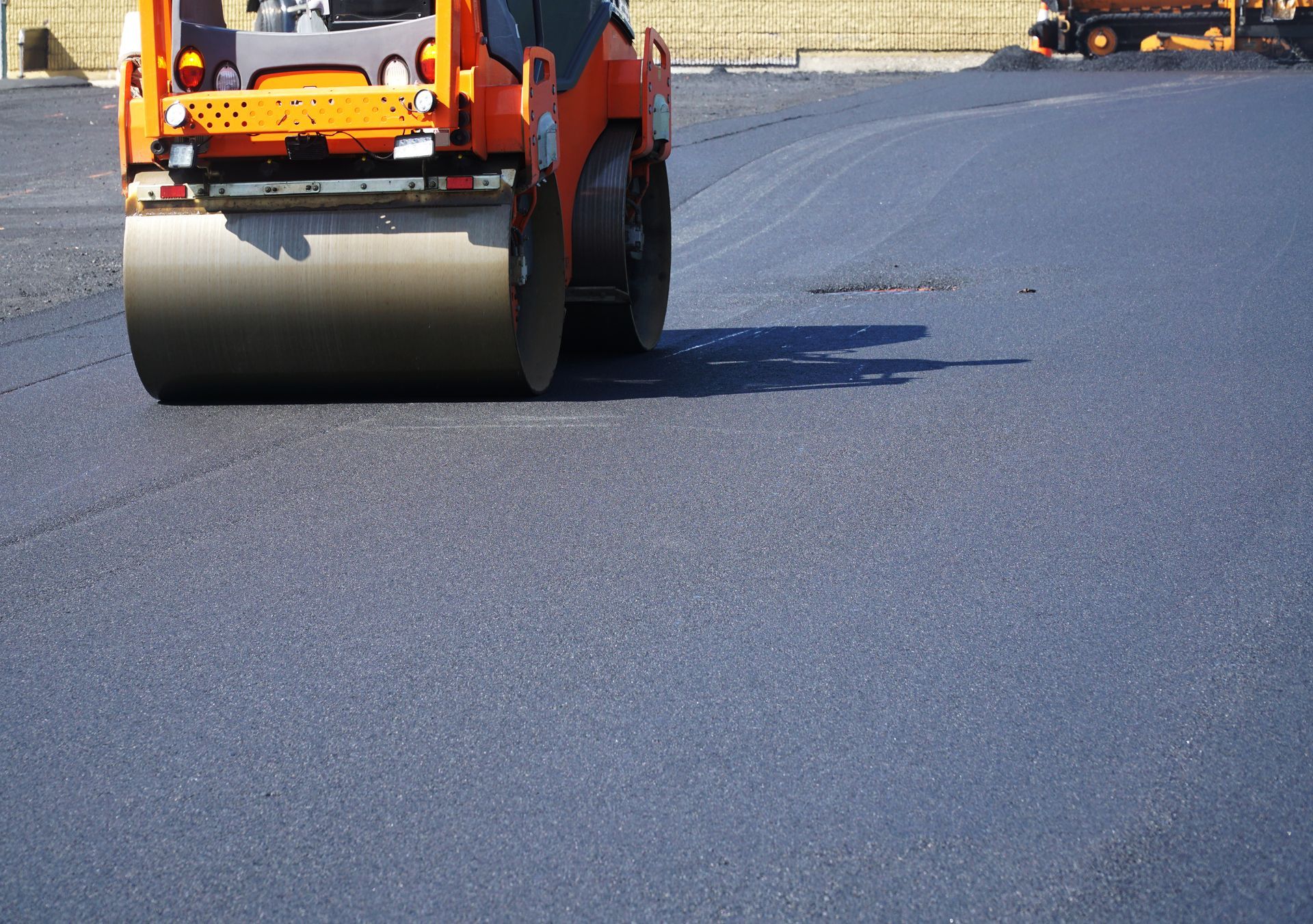 A compactor roller flattens a patch of asphalt on a road, under the sun. A compactor roller flattens a patch of asphalt on a road, under the sun.