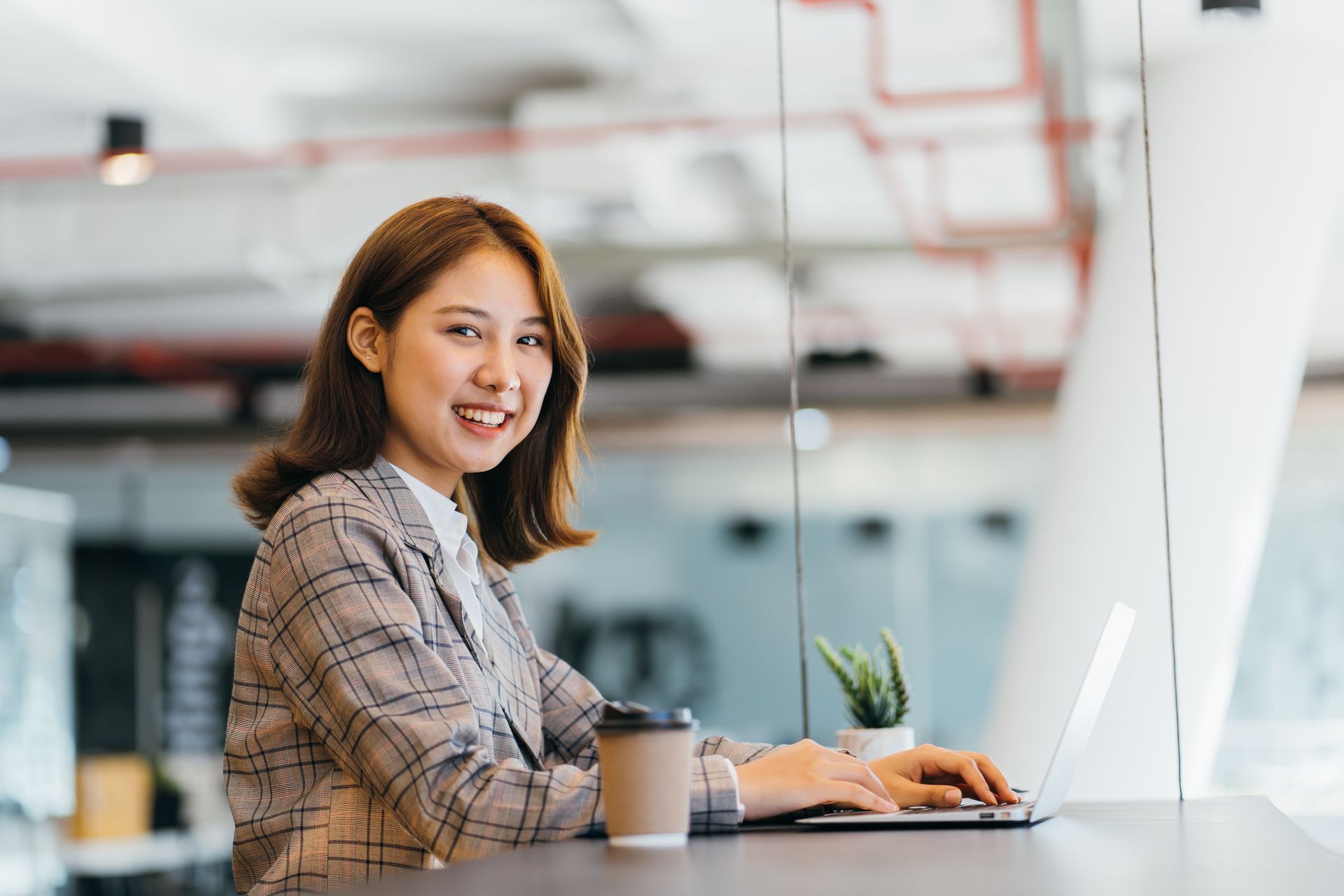 Woman in glasses at desk with video call on monitor, looking at graphs on tablet and paper.