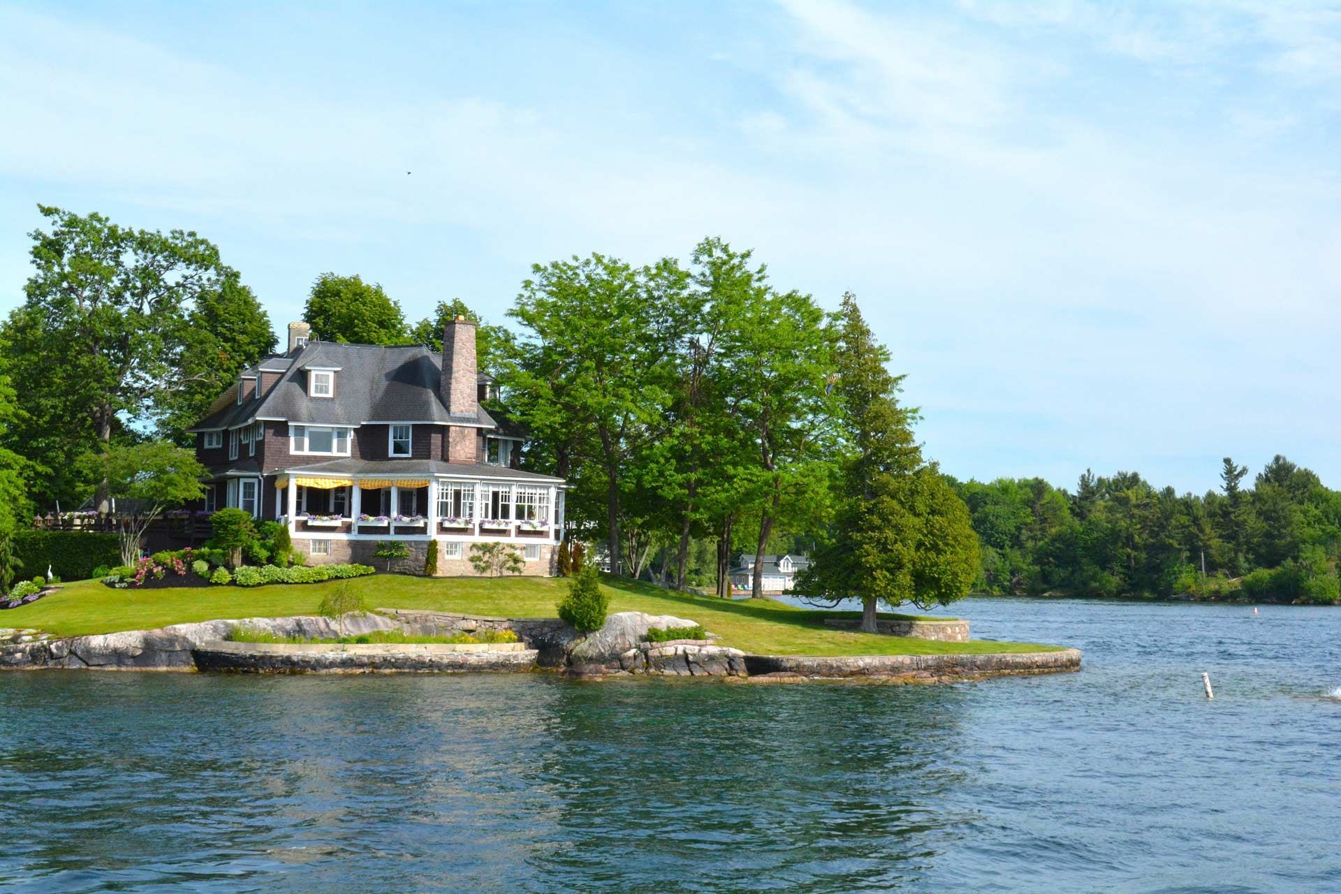 House on a grassy island peninsula, with trees and water. Blue sky.