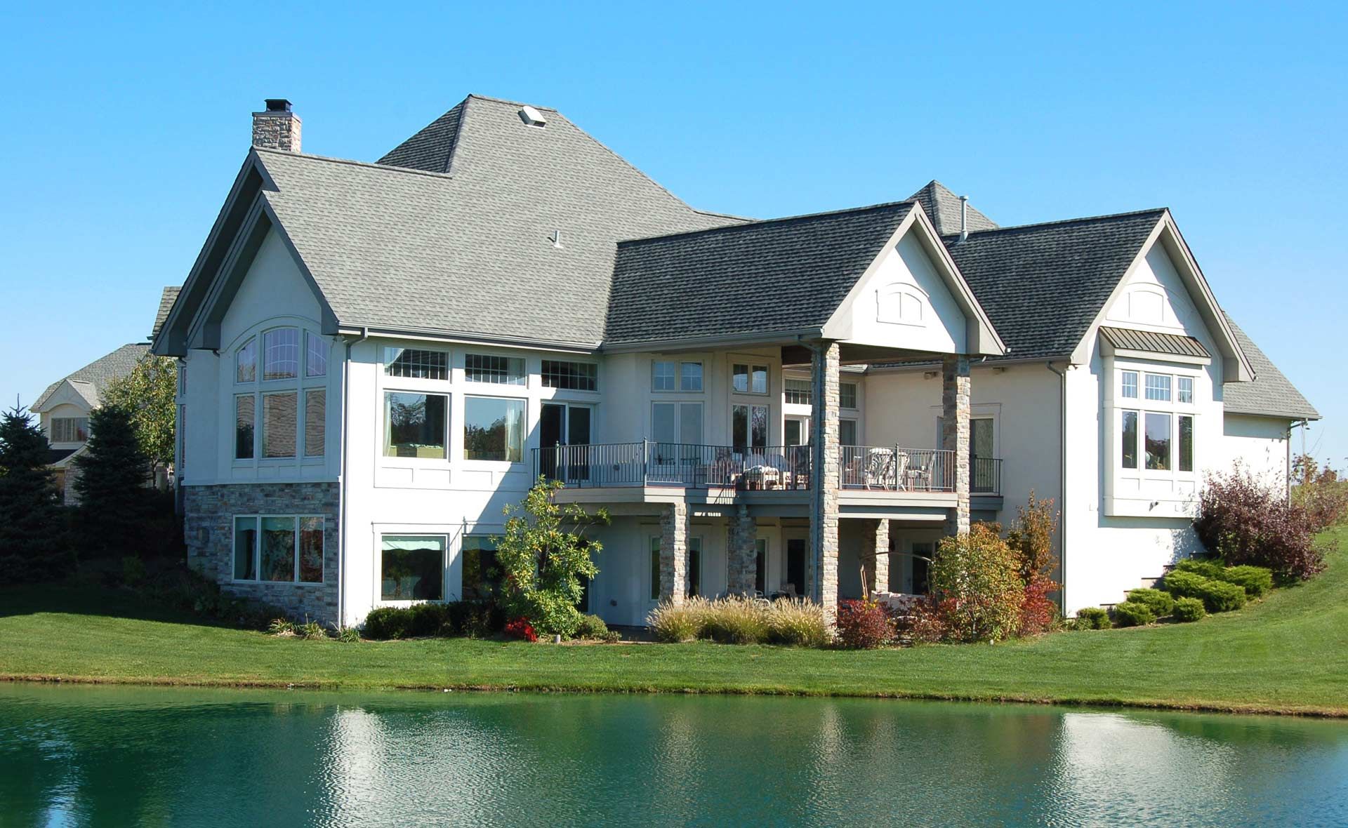 Large white house with gray roof and stone accents, overlooking a pond.