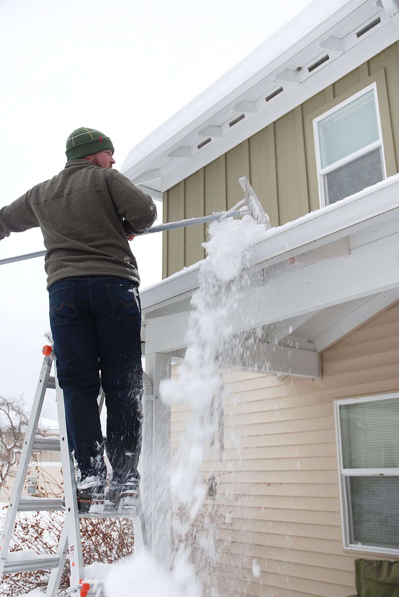 Man on ladder clearing snow from a gutter with a snow rake on a tan house with white trim.
