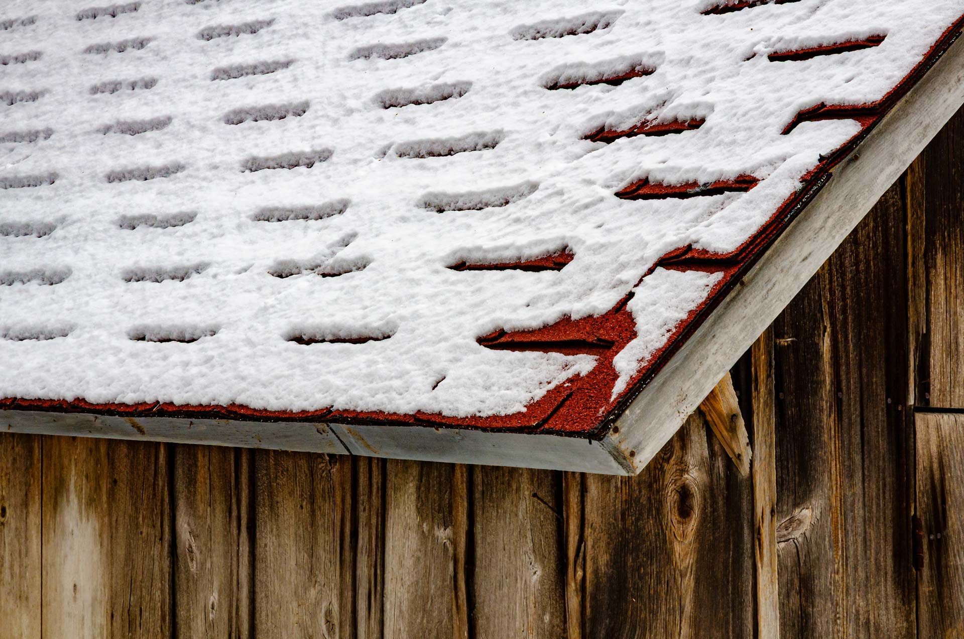 Snow-covered red shingle roof edge over weathered wooden siding.
