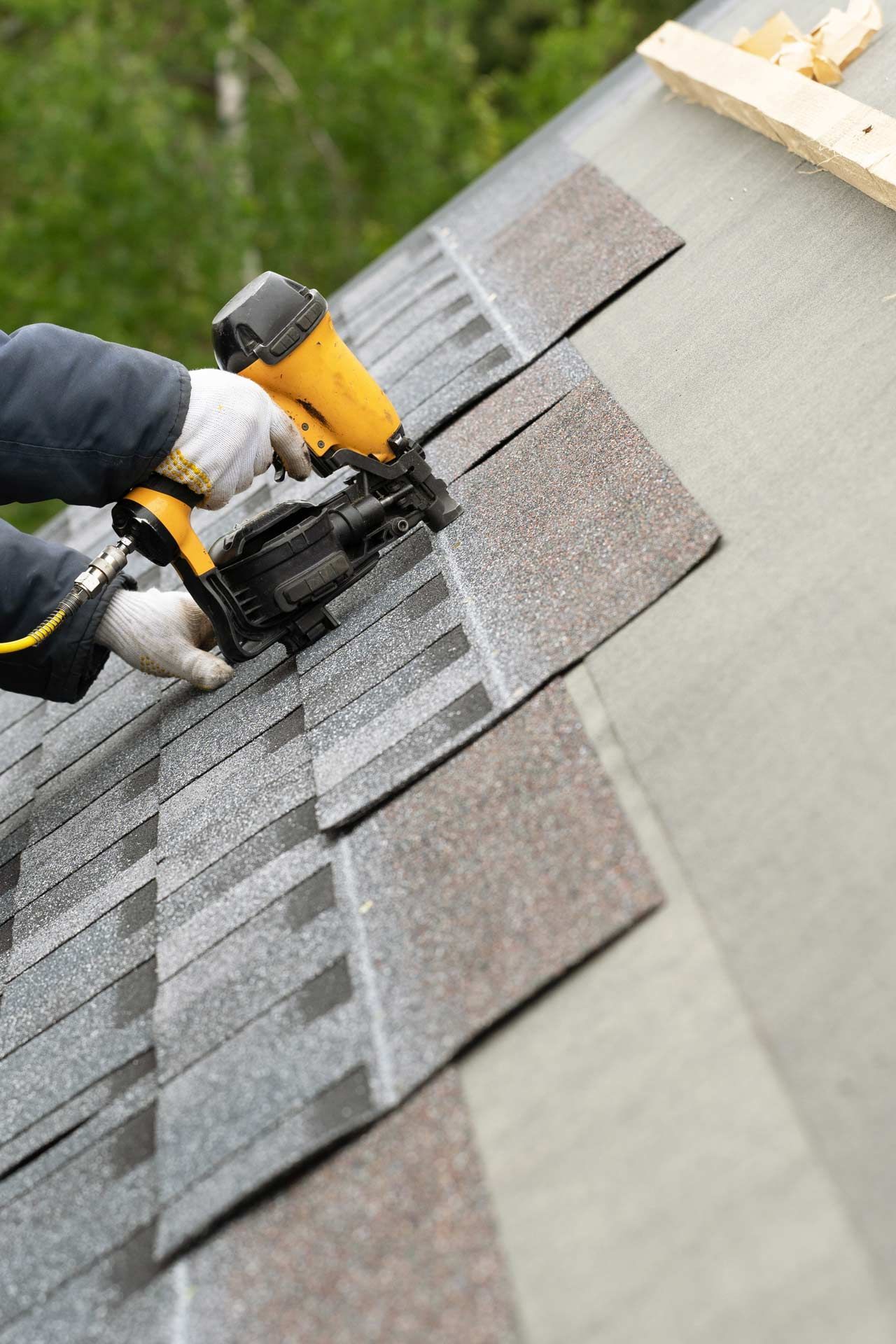 A worker using a nail gun to install roofing shingles on a slanted roof outdoors.