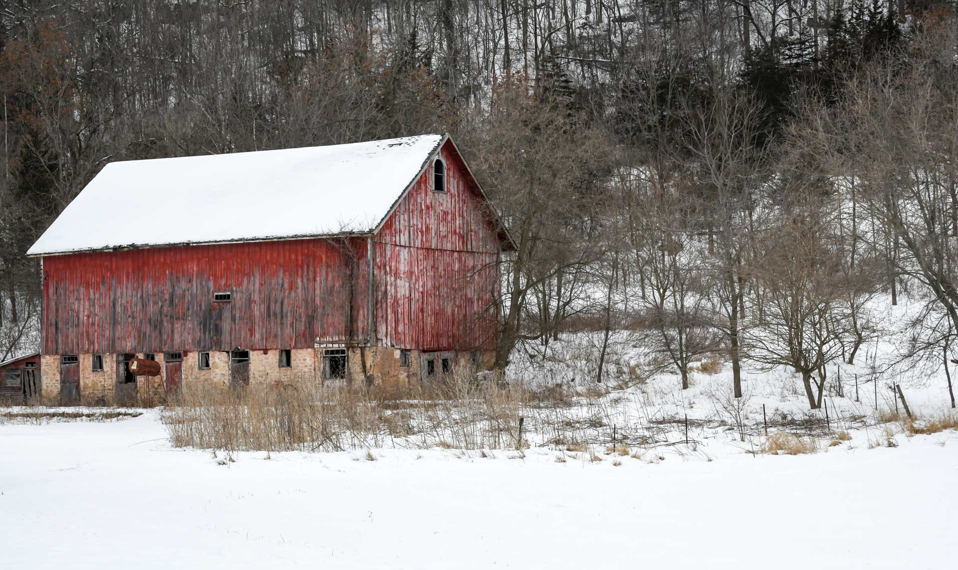 Red barn covered in snow, set in a snowy field with bare trees and a wooded hillside in the background.