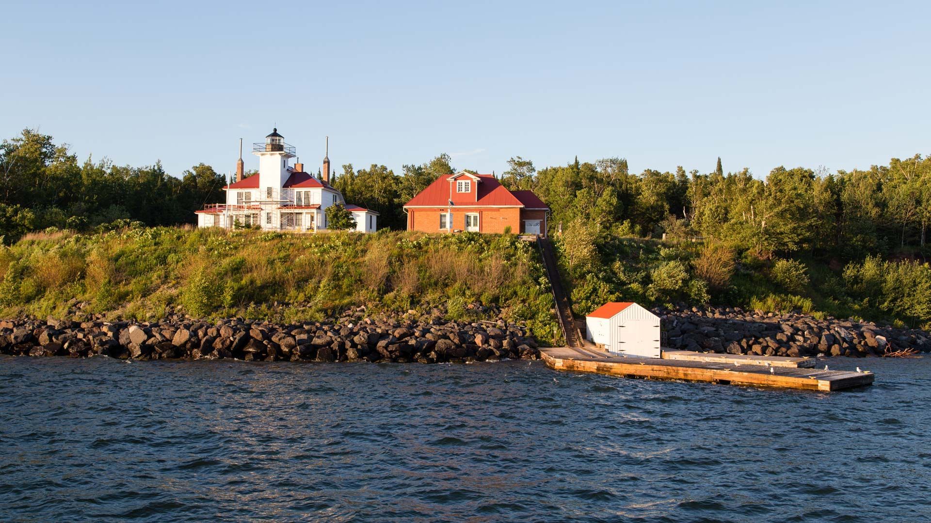 Lighthouse and buildings on a rocky shoreline with a small dock, blue water, and green trees.
