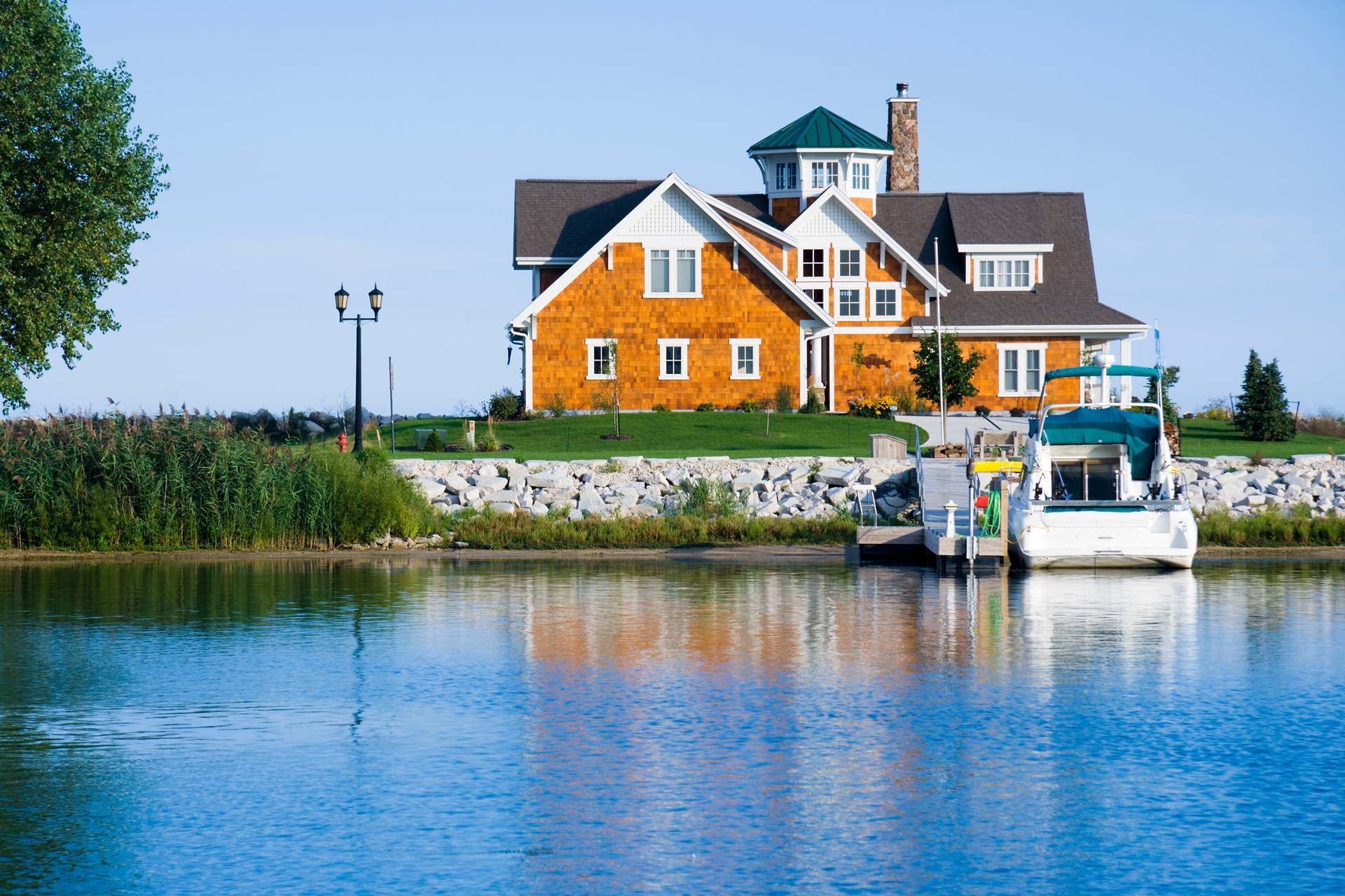 House with boats docked on a lake; orange siding, green roof, clear blue sky.