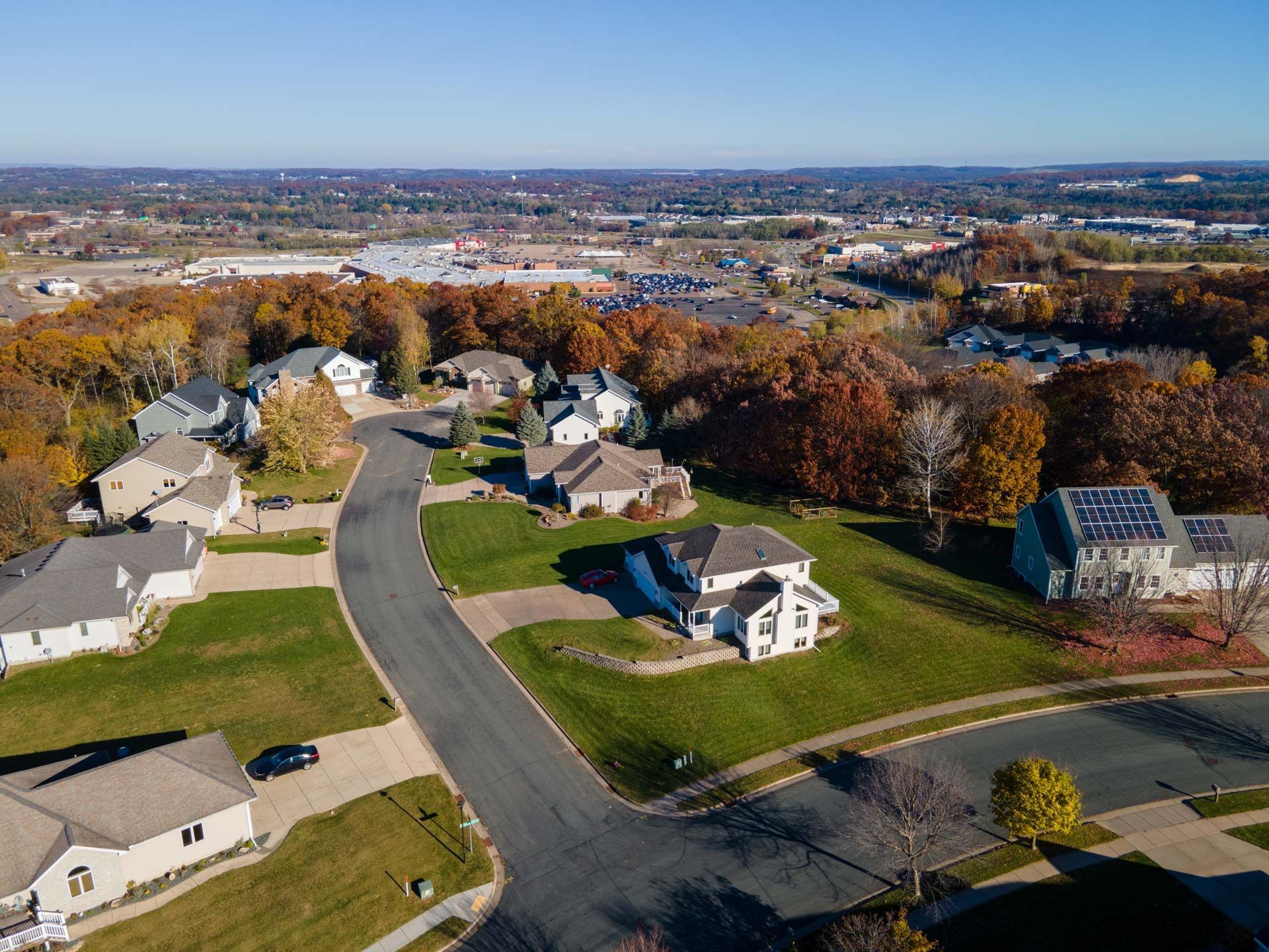 Aerial view of suburban houses on a curving street with autumn foliage and a distant commercial area under a blue sky.