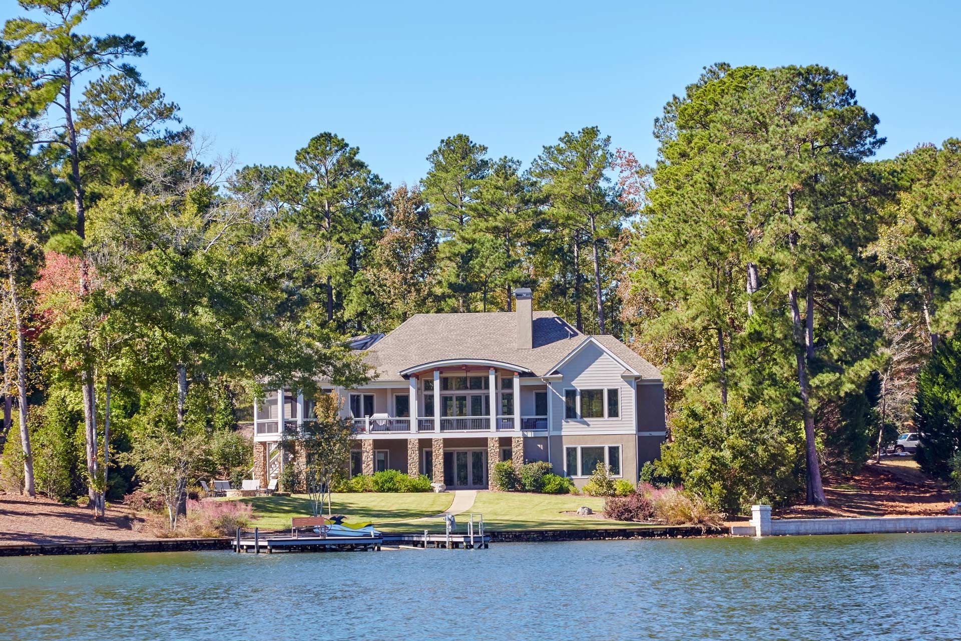 Two-story gray house with balconies on lakefront, surrounded by trees under blue sky.