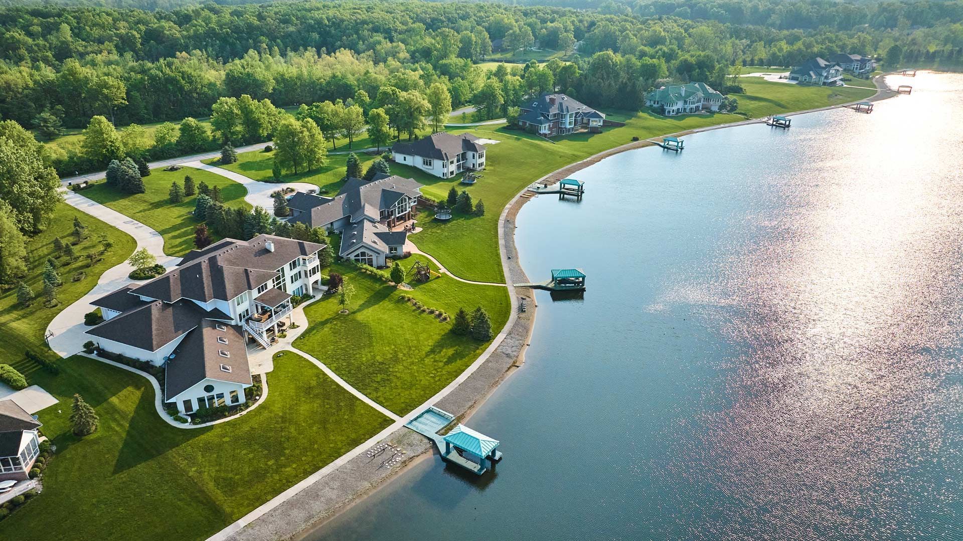 Lakeside luxury homes with private docks along a calm blue lake, surrounded by green lawns and trees.