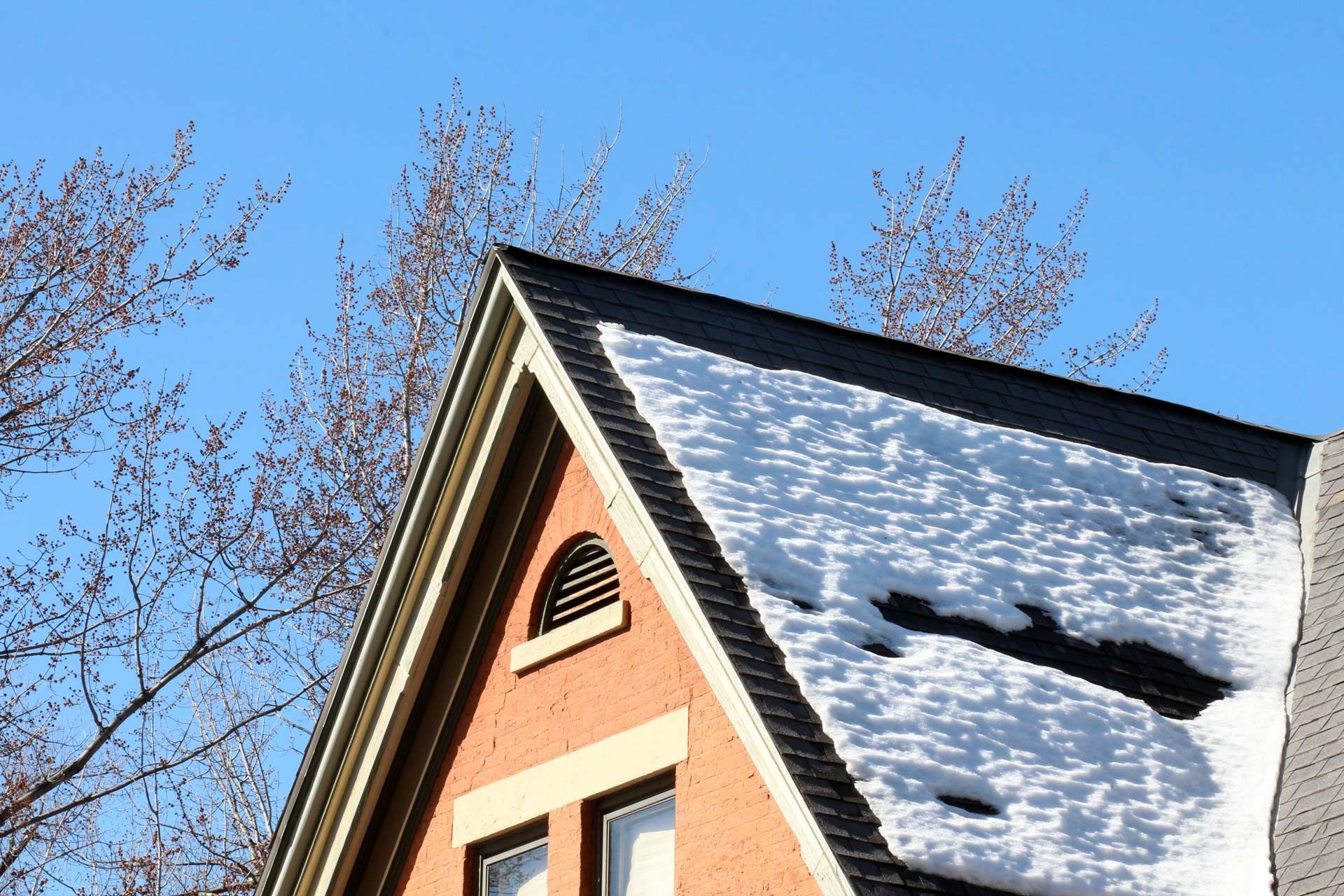Snow-covered roof of a brick house with bare tree branches against a clear blue sky.