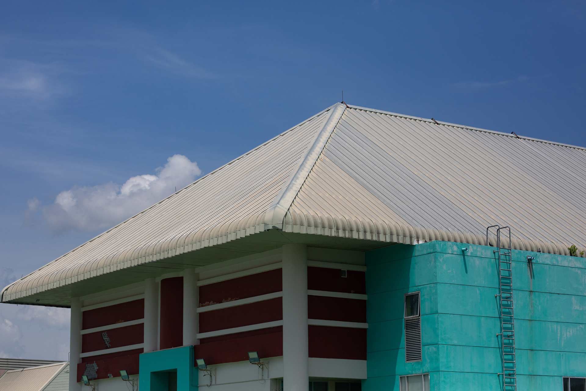 Building with a white roof, teal and red walls against a blue sky. A ladder is visible.