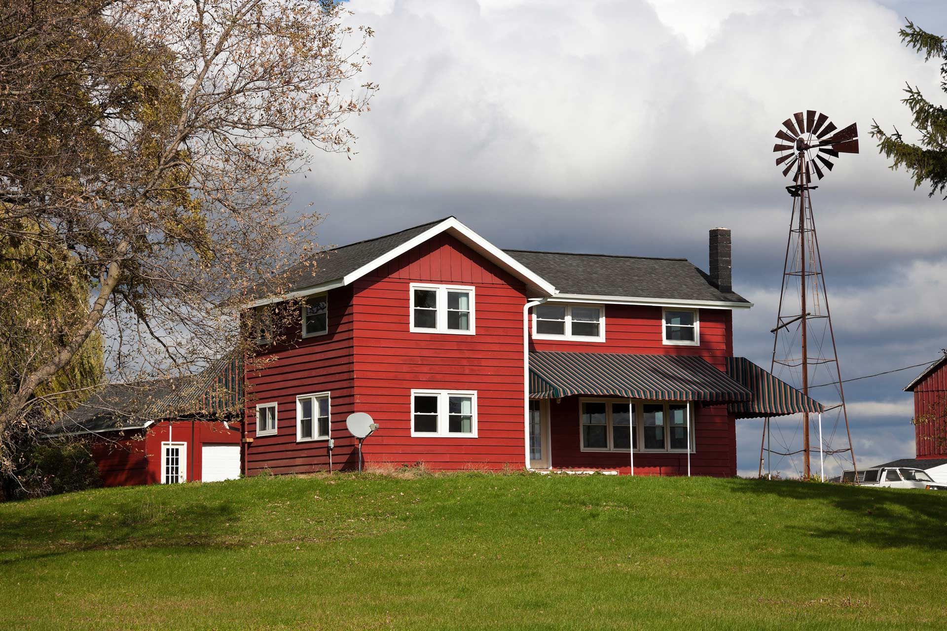 Red two-story farmhouse on a grassy hill, with a windmill and cloudy sky.